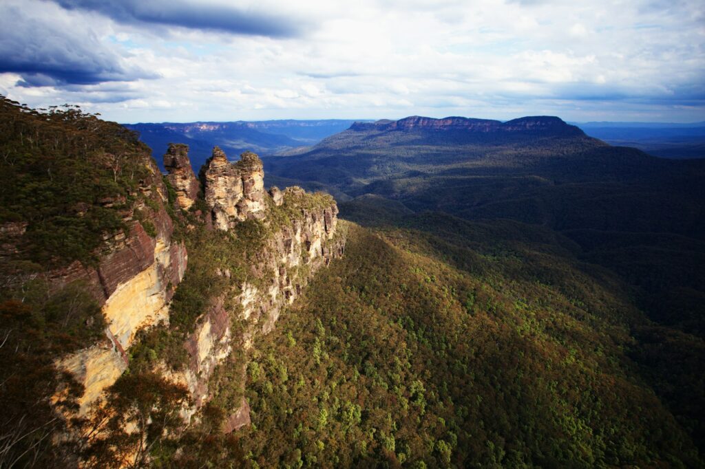 A rocky clifftop above a forested valley in the Blue Mountains, with the Three Sisters rock formation and distant ridges under a cloudy sky.