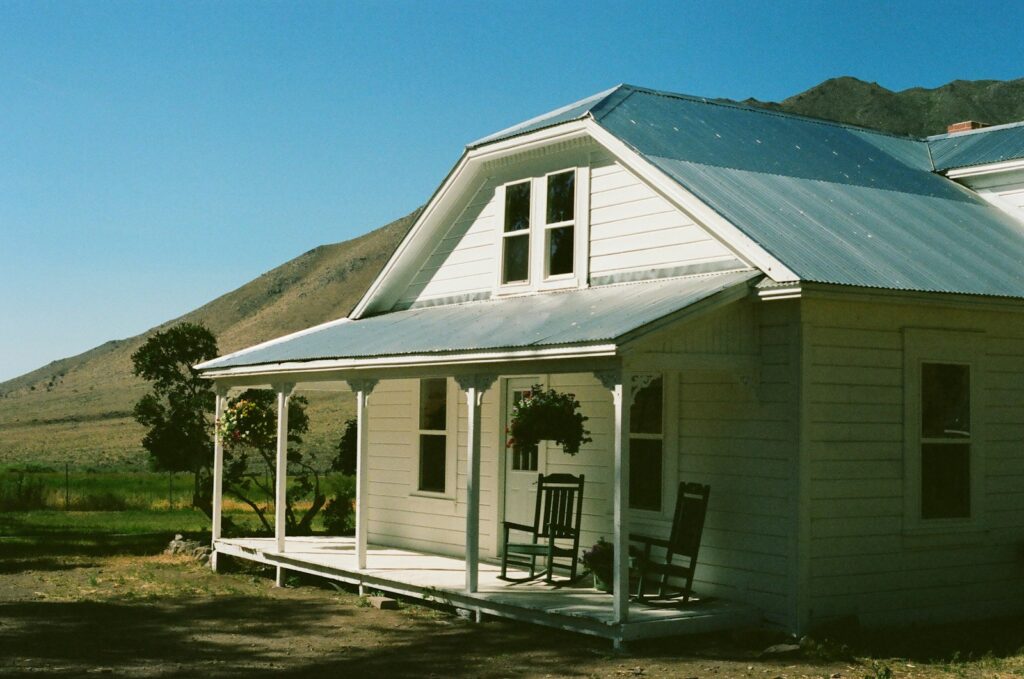 A white weatherboard farmhouse with a veranda and rocking chairs, green hills behind.